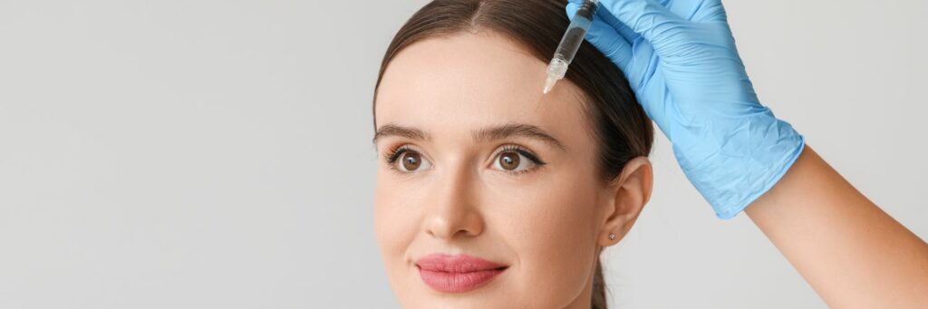 Eine Person mit braunem Haar erhält von einer behandschuhten Hand, die eine Spritze hält, eine Injektion in die Stirn, möglicherweise für ein Brauenlifting.