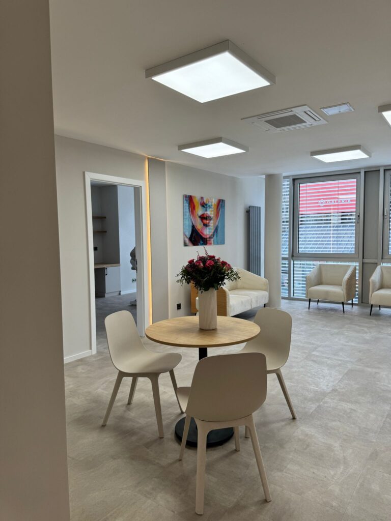 Ein moderner büroraum mit einem runden tisch, vier stühlen und einer blumenvase. helle deckenbeleuchtung und farbenfrohe wandkunst. im hintergrund stehen weiße sessel neben großen fenstern.