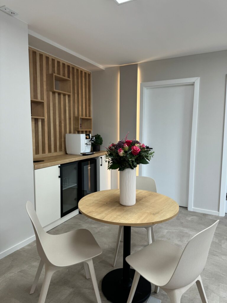Kleine küchenzeile mit holzelementen, schwarzem minikühlschrank und kaffeemaschine. runder tisch mit drei stühlen und einer vase mit rosa blumen in der mitte. weiße wände und eine tür im hintergrund.