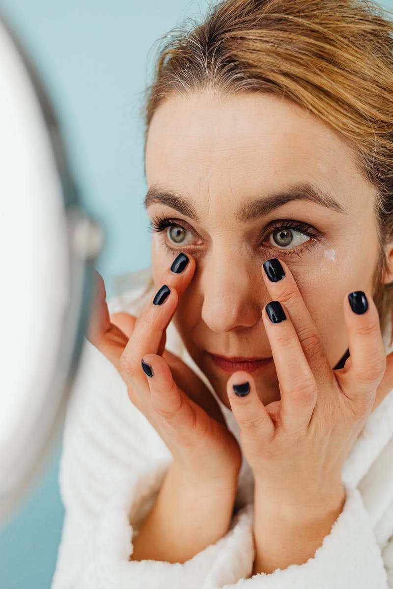 A woman in a white robe applying under-eye cream while looking in the mirror.