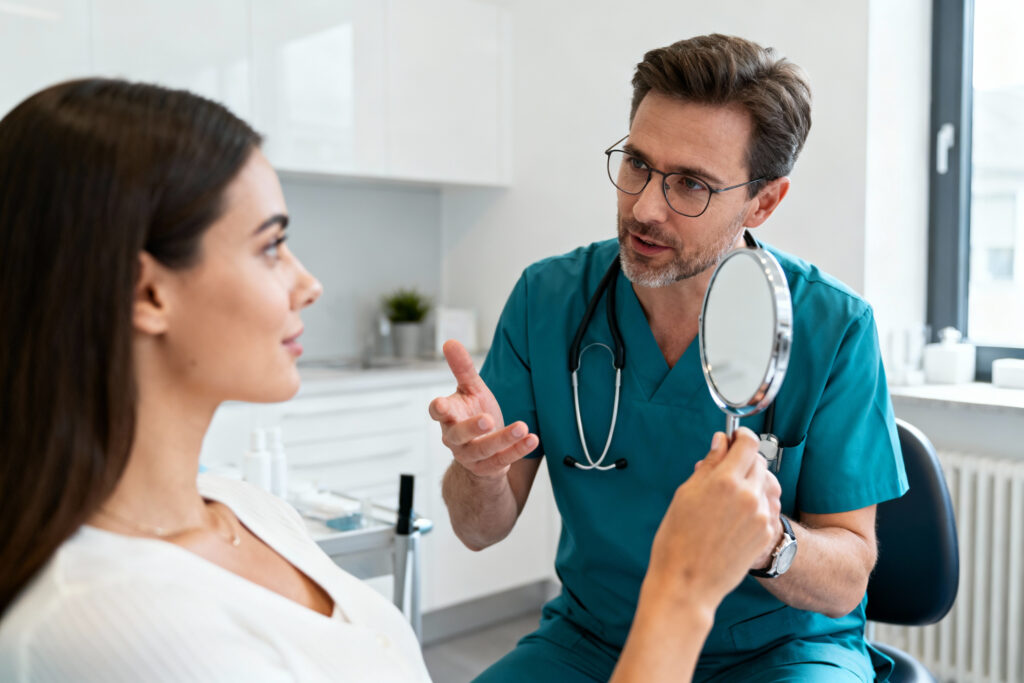 Ein Arzt im Kittel spricht mit einer Frau, die einen Spiegel hält, während einer Konsultation in einer Arztpraxis.